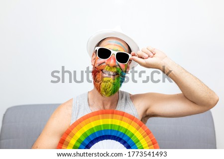 white man smiling and in white hat and gray T-shirt with face and beard painted colors of lgtbi