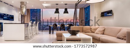 Panorama of luxury decorated living room with kitchen in apartment with big window wall during sunset Royalty-Free Stock Photo #1773572987