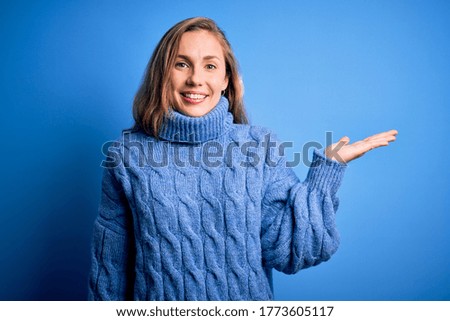 Young beautiful blonde woman wearing casual turtleneck sweater over blue background smiling cheerful presenting and pointing with palm of hand looking at the camera.