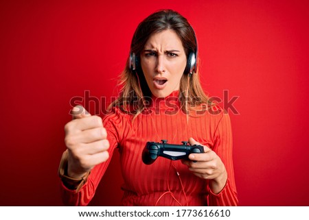 Young beautiful brunette gamer woman playing video game using joystic and headphones annoyed and frustrated shouting with anger, crazy and yelling with raised hand, anger concept