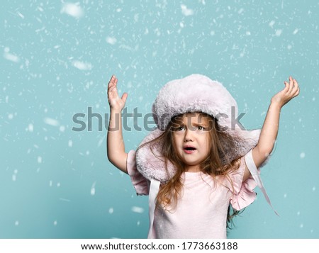 Little blond kid with long hair, dressed in a pink blouse, a plush hat with earflaps. She raised her hands, looking confused, posing on a blue background in snowfall.
