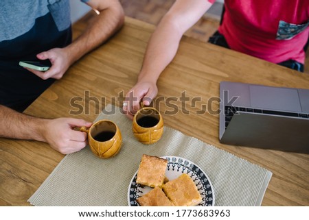 Stock photo from top of a wooden table. There is a laptop, a mobile phone, two mugs of coffee and a plate of biscuits. There are arms of two caucasian people.