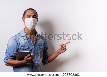 Portrait of a young Latino man on a white background wearing a medical mask holding a tablet and pointing his finger towards the empty space on the left side. Copy space. Quarantine concept.
