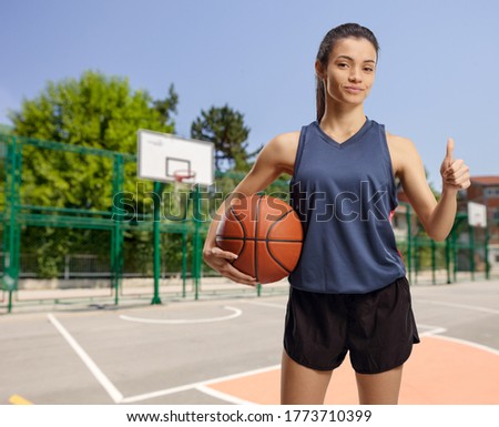 Young female with a basketball standing on an outdoor court and showing a thumb up sign 