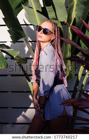 Young europian stylish woman with pink purple braids in gray long shirt, accessories, sunglasses, black waist bag trendy fashion watch clock on hand, on tropical background and white wooden wall.