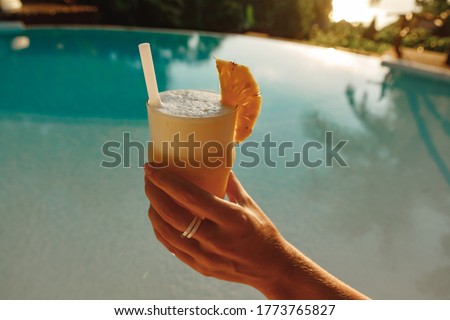 Glass of pineapple juice in woman hands on background of the swimming pool. Refreshment drink, summer concept. Young woman by the sunny pool holding a cocktail in her hand