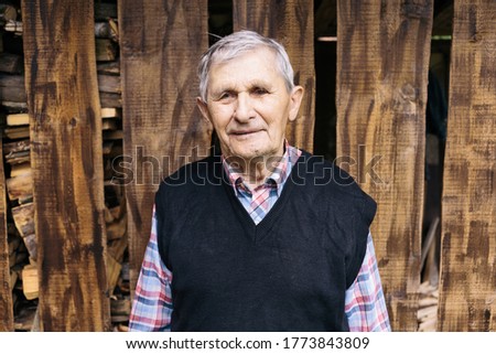 Happy old grandfather smiling and standing on the background of a wooden fence in the yard in the village. The older generation