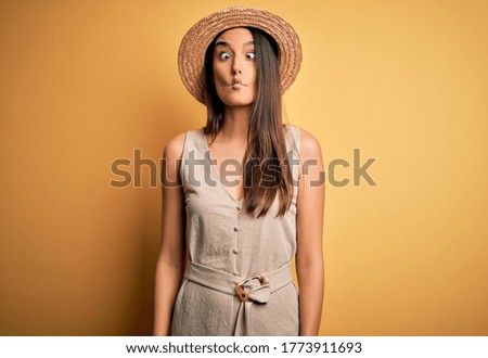Young beautiful brunette woman on vacation wearing casual dress and hat making fish face with lips, crazy and comical gesture. Funny expression.