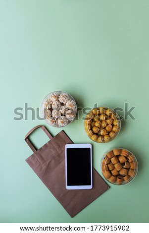 Blank screen tablet on brown paper bag and jars of assorted cookies with light green background. Online business concept.
