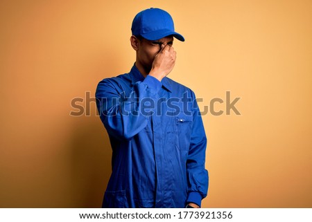 Young african american mechanic man wearing blue uniform and cap over yellow background tired rubbing nose and eyes feeling fatigue and headache. Stress and frustration concept.