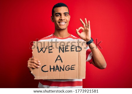 Young handsome african american activist man asking for change holding banner with message doing ok sign with fingers, excellent symbol