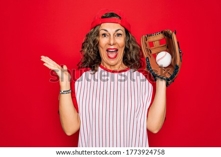 Middle age senior woman wearing baseball equiment, ball and glove over red isolated background very happy and excited, winner expression celebrating victory screaming with big smile and raised hands