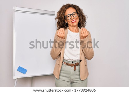 Middle age senior business woman standing on seminar presentation by magnectic blackboard doing money gesture with hands, asking for salary payment, millionaire business