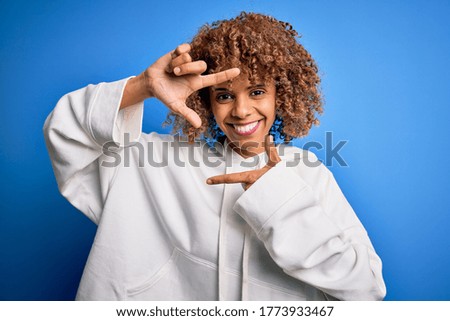 Beautiful african american sporty woman wearing casual sweatshirt over blue background smiling making frame with hands and fingers with happy face. Creativity and photography concept.