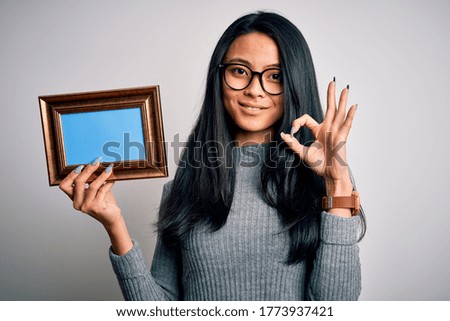 Young beautiful chinese woman holding vintage frame over isolated white background doing ok sign with fingers, excellent symbol