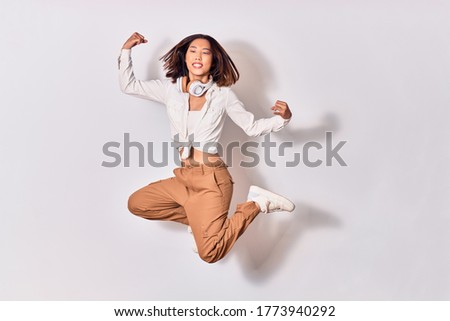 Young beautiful chinese woman smiling happy. Jumping with smile on face listening to music using headphones doing strong gesture with arms and fists up over white background