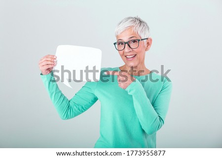Happy mature woman in plain blue long sleeve t-shirt holding empty speech bubble isolated on background. Woman showing sign speech bubble banner looking happy and pointing her finger on it
