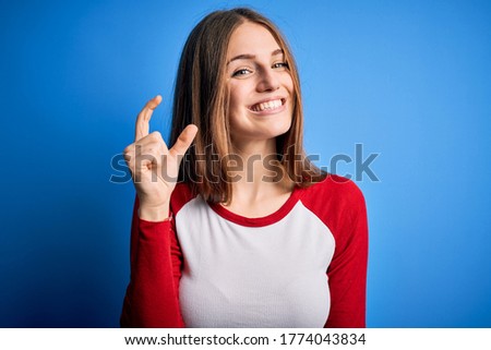 Young beautiful redhead woman wearing casual t-shirt over isolated blue background smiling and confident gesturing with hand doing small size sign with fingers looking and the camera. Measure concept.