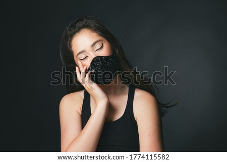 Glamorous trendy asian girl in fashionable black face shield. Covid-19 quarantine fashion style. Stylish graphic makeup. Beautiful designer beaded accessory. closed eyes headache. Blinged out face 