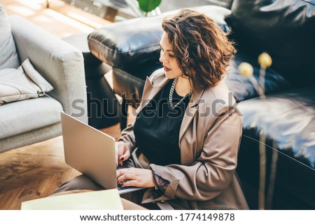 Caucasian brunette woman sitting near sofa in living room and using laptop for freelance job.