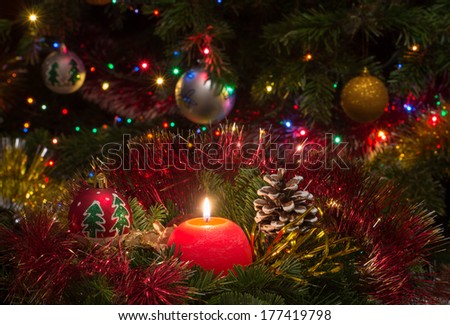 A red candle surrounded by pine branches with Christmas lights,Christmas balls, tree and Santa Clouse in the background.