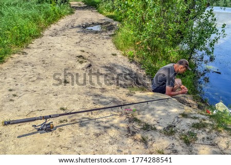 Fisherman puts bait on fishing rod hook - top view. Closeup fishing rod lies on the ground and a young adult man sits on the shore against the backdrop of the water