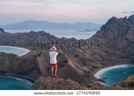 Young Asian man traveller standing and taking picture on top of Padar island in a morning sunrise, Komodo national park in Flores island in summer season, Indonesia, Asia