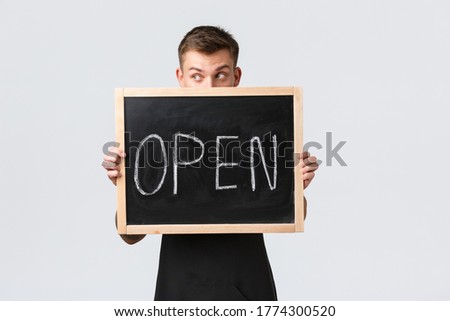 Small retail business owners, cafe and restaurant employees concept. Silly handsome barista, salesman looking away, holding open sign near face, standing white background