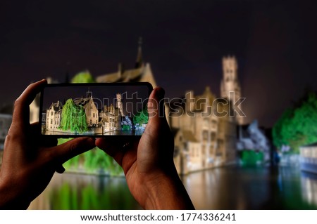 Tourist taking photo of canal of Bruges, tower and historic buildings at night, Bruges, Belgium. Man holding phone and taking picture.