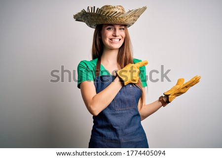 Young beautiful redhead farmer woman wearing apron and hat over white background amazed and smiling to the camera while presenting with hand and pointing with finger.
