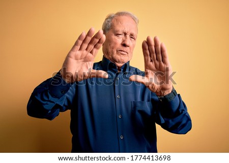 Grey haired senior man wearing casual blue shirt standing over yellow background doing frame using hands palms and fingers, camera perspective