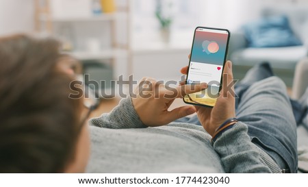 Man at Home Lying on a Couch using Smartphone, Scrolling Social Media Feed, Checking News, Friends Activity. Guy Using Mobile Phone, Internet Social Networks Browsing. Royalty-Free Stock Photo #1774423040