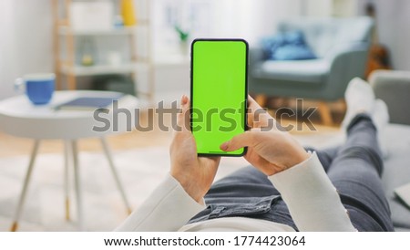 Woman at Home Lying on a Couch using Smartphone with Green Mock-up Screen, Doing Swiping, Scrolling Gestures. Girl Using Mobile Phone, Internet Social Networks Browsing. Point of View Shot.