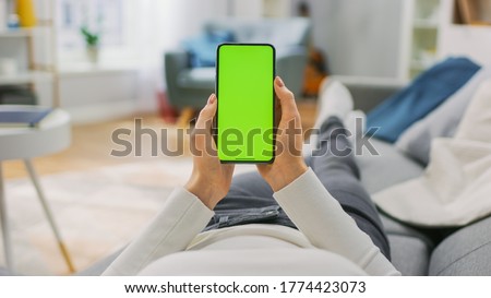Young Woman at Home Uses Green Mock-up Screen Smartphone. She's Sitting On a Couch in His Cozy Living Room. Point of View Shot.