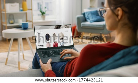 Young Woman at Home Using Laptop Computer for Browsing Through Online Retail Shopping Site. She's Sitting On a Couch in His Cozy Living Room. Over the Shoulder Camera Shot Royalty-Free Stock Photo #1774423121
