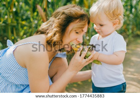 Picture of charming young caucasian woman with short hair in light blue dress relaxes with her little male baby with short fair hair in blue t-shirt and blue shorts in cornfield.