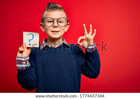 Young little caucasian kid wearing glasses holding paper note with question mark sign doing ok sign with fingers, excellent symbol