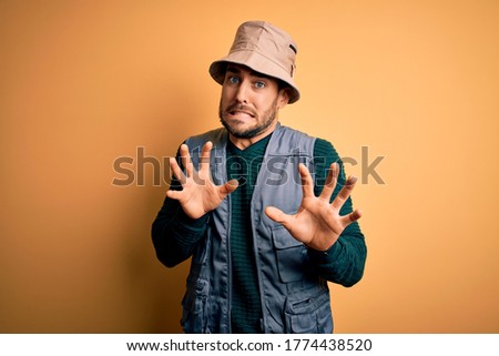 Handsome tourist man with beard on vacation wearing explorer hat over yellow background disgusted expression, displeased and fearful doing disgust face because aversion reaction.