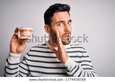 Young handsome man with beard holding plastic denture teeth over white background hand on mouth telling secret rumor, whispering malicious talk conversation