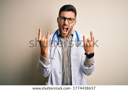 Young doctor man wearing glasses, medical white robe and stethoscope over isolated background shouting with crazy expression doing rock symbol with hands up. Music star. Heavy concept.