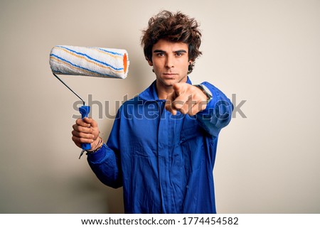 Young handsome painter man holding roller wearing uniform over isolated white background pointing with finger to the camera and to you, hand sign, positive and confident gesture from the front