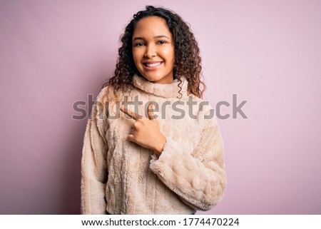 Young beautiful woman with curly hair wearing casual sweater standing over pink background cheerful with a smile on face pointing with hand and finger up to the side with happy and natural expression