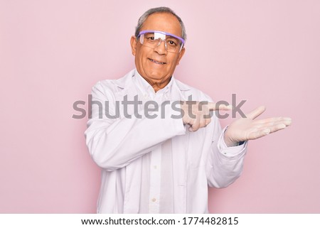 Senior grey-haired scientist man wearing glasses and coat over isolated pink background amazed and smiling to the camera while presenting with hand and pointing with finger.