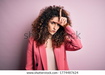 Young beautiful businesswoman with curly hair and piercing wearing elegant jacket making fun of people with fingers on forehead doing loser gesture mocking and insulting.