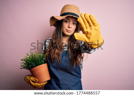 Young beautiful brunette farmer woman wearing apron and hat holding pot with plants with open hand doing stop sign with serious and confident expression, defense gesture