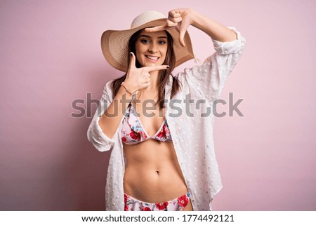 Young beautiful woman on vacation wearing bikini and summer hat over pink background smiling making frame with hands and fingers with happy face. Creativity and photography concept.