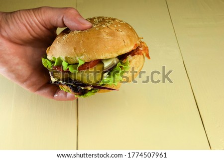 Against the background of a beige wooden table, the hand holds a sandwich with meat, cheese, tomatoes, onions, cucumbers and salad