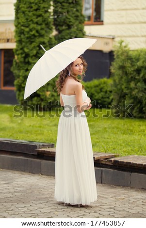 Beautiful happy bride in white dress with decorative umbrella against background green nature. In a summer park
