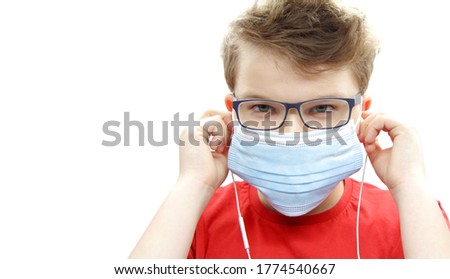 Portrait of a boy in a surgical medical blue mask against covid-19 coronavirus. A teenager in white wired headphones, glasses, and a red t-shirt listens to music on a white background. Copyspace.