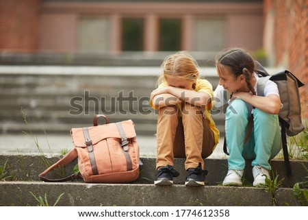 Full length portrait of teenage schoolgirl crying while sitting on stairs outdoors with smiling friend comforting her, copy space Royalty-Free Stock Photo #1774612358